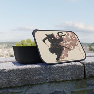 May include: A black lunchbox featuring a ninja cat illustration. The cat is shown in action, holding swords, against a smoky brown backdrop. The open lunchbox reveals a black container filled with green grapes.