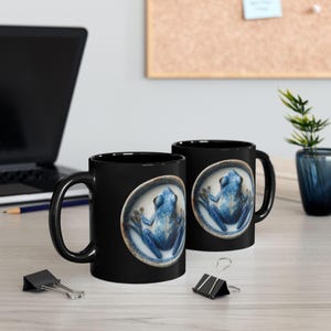 May include: Two black ceramic mugs with a blue frog design. The frog is centered on a white oval, with a detailed illustration of the frog's features. The mugs are on a light-colored wooden surface.
