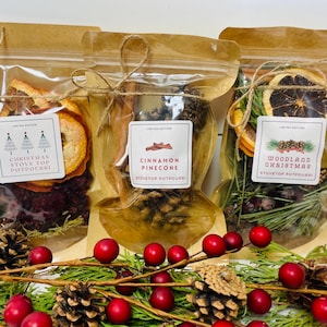 May include: Three clear bags of stovetop potpourri, each with a different scent: Christmas, Cinnamon Pinecone, and Woodland Christmas. The bags are tied with twine and have labels. The image includes pinecones, red berries, and greenery.