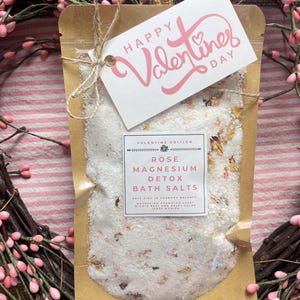 May include: A brown paper pouch with a white tag that says "Happy Valentine's Day" and contains white bath salts with pink rose petals. The pouch is labeled "Valentine's Edition Rose Magnesium Detox Bath Salts".