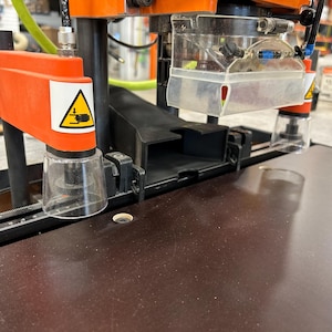 Puede incluir: Primer plano de una máquina para trabajar la madera naranja y negra. La máquina tiene un protector de plástico transparente y una etiqueta de advertencia. La superficie de trabajo es de color marrón oscuro. La máquina está diseñada para cortar y dar forma a la madera con precisión.
