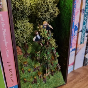 May include: A miniature forest scene with two small figures climbing a tree, set inside a bookend. The bookend is made of wood and has a green background. The forest scene is made of moss and small trees. The bookend is on a shelf with other books.
