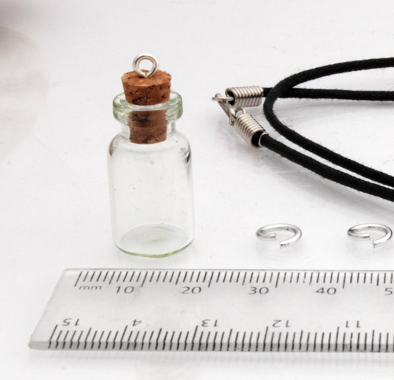 A small jar shaped glass vial with a cork lid and metal loop stands on a white background beside a ruler for scale. A black necklace cord is behind, all sat on a white background