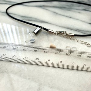 A miniature glass vial with silver coloured caps shown on a white marble slab with a ruler for scale. The vial looks like an amphora, rounded with a pointed base. 2 jump rings, a rubber bung and a black pendant cord seen in the background.