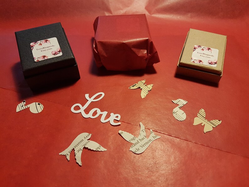 May include: Three small gift boxes wrapped in brown paper, black paper, and red tissue paper. The boxes are on a red surface with paper cutouts of birds, butterflies, and musical notes. The word "Love" is written in white paper.