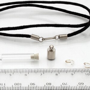 A miniature glass test tube shaped vial with silver coloured cap is shown on a white marble slab with a ruler for scale. 2 jump rings, a rubber bung and a black pendant cord seen in the background.