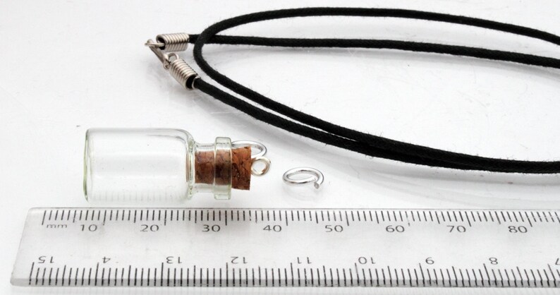 A small jar shaped glass vial with a cork lid and metal loop lays on its side on a white background beside a ruler for scale. A black necklace cord is behind, all sat on a white background