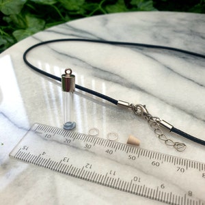 A miniature glass test tube shaped vial with silver coloured cap is shown on a white marble slab with a ruler for scale. 2 jump rings, a rubber bung and a black pendant cord seen in the background.