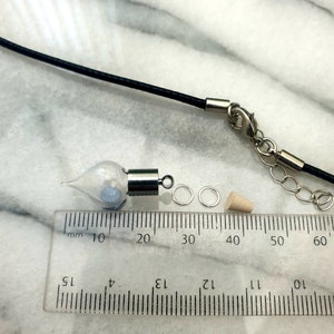 A miniature glass vial with silver coloured caps shown on a white marble slab with a ruler for scale. The vial looks like an amphora, rounded with a pointed base. 2 jump rings, a rubber bung and a black pendant cord seen in the background.