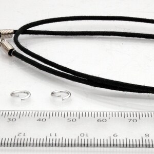 A small jar shaped glass vial with a cork lid and metal loop stands on a white background beside a ruler for scale. A black necklace cord is behind, all sat on a white background