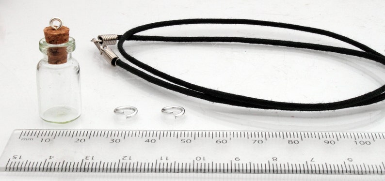 A small jar shaped glass vial with a cork lid and metal loop stands on a white background beside a ruler for scale. A black necklace cord is behind, all sat on a white background