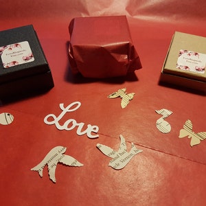 May include: Three small gift boxes wrapped in brown paper, black paper, and red tissue paper. The boxes are on a red surface with paper cutouts of birds, butterflies, and musical notes. The word "Love" is written in white paper.