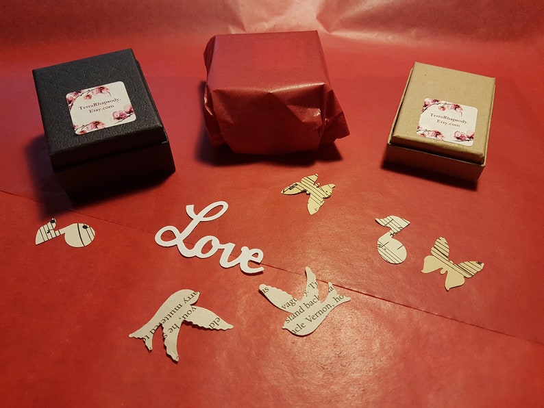 May include: Three small gift boxes wrapped in brown paper, black paper, and red tissue paper. The boxes are on a red surface with paper cutouts of birds, butterflies, and musical notes. The word "Love" is written in white paper.