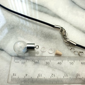 A miniature glass globe shaped vial with silver coloured cap is shown on a white marble slab with a ruler for scale. 2 jump rings, a rubber bung and a black pendant cord seen in the background.