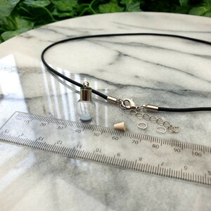 A miniature glass globe shaped vial with silver coloured cap is shown on a white marble slab with a ruler for scale. 2 jump rings, a rubber bung and a black pendant cord seen in the background.
