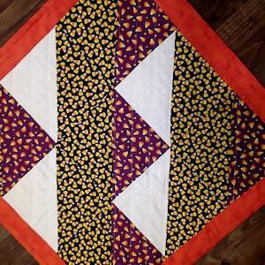 May include: A Halloween-themed quilt with a patchwork design featuring orange, white, purple, and black fabrics. The quilt is made with a candy corn print fabric and a white fabric with small white dots. The quilt has an orange border.