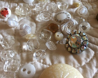 May include: A collection of vintage buttons in various colors and styles, including white, clear, and gold. Some buttons have intricate designs, while others are plain. The buttons are arranged on a white fabric background.