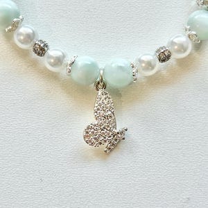 May include: A delicate white and light blue beaded necklace with a silver butterfly charm. The butterfly charm is adorned with sparkling rhinestones.