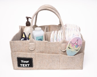 May include: Beige felt diaper bag with multiple pockets and compartments. The bag has a black label that says "Your Text". The bag is filled with baby supplies, including diapers, lotion, and a unicorn-shaped hairbrush.