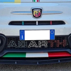 Op de afbeelding: Voorkant van een witte auto met het woord "ABARTH" in de grille. Voorzien van een rode sleepband, Italiaanse vlagkleuren en het Abarth-logo. De auto heeft een kentekenplaat.