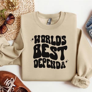 Op de afbeelding: Een beige sweatshirt met de tekst "WORLD'S BEST DEPENDA" in zwarte, retro-stijl letters.