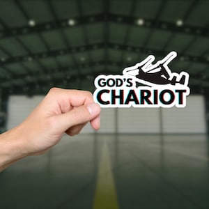 May include: A white sticker with the words "GOD'S CHARIOT" in black and red, featuring a black helicopter silhouette. The sticker is held against a blurred background of a building interior.