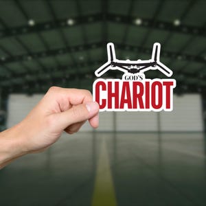May include: A white sticker with a black graphic of an aircraft and the words "GOD'S CHARIOT" in red. The sticker is held by a hand against a blurred background of a building interior.