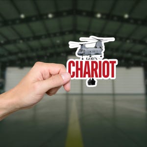 May include: A sticker featuring a gray helicopter design above the words "GOD'S CHARIOT" in red, outlined in white. The sticker is held in a person's hand against a blurred background of a large building.