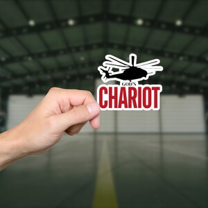 May include: A sticker featuring a black helicopter silhouette above the words "GOD'S CHARIOT" in red, outlined in white. The sticker is held by a hand against a blurred background.