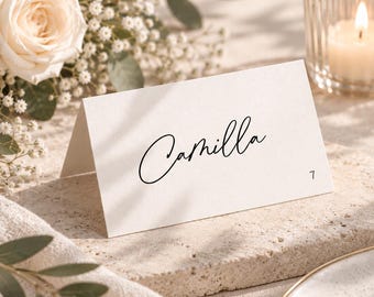 Può includere: Un segnaposto bianco piegato con il nome Camilla scritto in corsivo nero e il numero 7 stampato nell'angolo in basso a destra. Il cartoncino è realizzato in carta spessa con una finitura opaca pulita e una piega centrale netta e precisa.