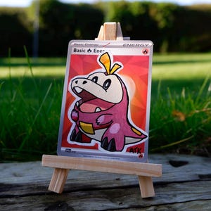 May include: A hand-painted Pokémon trading card depicting a pink and white crocodile-like creature with yellow details. The card is presented on a miniature wooden easel, set against a backdrop of green grass and a blurred background.