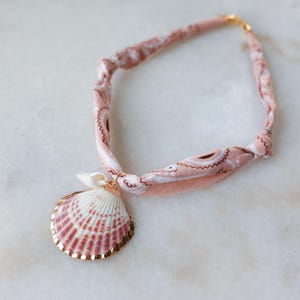 May include: A seashell pendant necklace. The necklace features a pink and white paisley fabric band with a gold-colored clasp. The pendant is a scallop shell with pink and white stripes and a gold-colored trim. The necklace is a beach-themed accessory.
