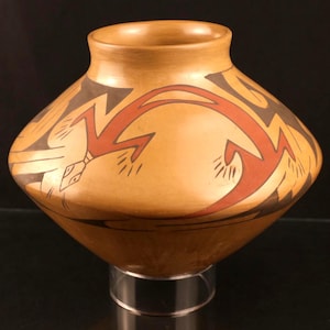 May include: A tan-colored pottery vase with a rounded body and a narrow neck. The vase features a geometric design in red and black, with a stylized bird or arrow motif. The vase is displayed on a clear stand.