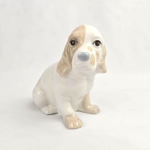 May include: A ceramic figurine of a dog in a seated position. The dog has a white body with tan markings on its head and ears. It has brown eyes and a light blue nose. The figurine is a decorative item.