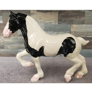May include: A ceramic figurine of a horse foal in a walking pose. The foal is primarily white with black markings on its head and body. The hooves and nose are pink.