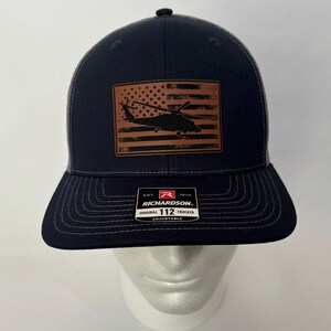 Puede incluir: Una gorra de camionero azul marino y gris con un parche marrón que presenta una bandera estadounidense desgastada con una silueta negra de un helicóptero. La gorra tiene una correa ajustable negra con una etiqueta roja y blanca que dice "Richardson 112 Original Trucker Adjustable".