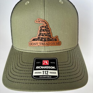 Puede incluir: Gorra Trucker Richardson Original 112 verde oliva con parte trasera de malla negra. Parche de cuero marrón con una serpiente enroscada y el texto "DON'T TREAD ON ME". Ajustable.
