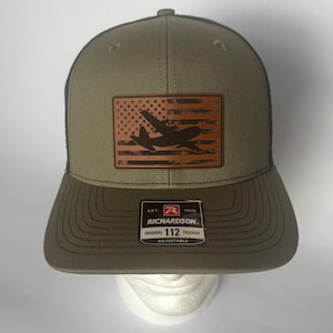 May include: Olive green and black trucker hat with a brown leather patch featuring an airplane silhouette over an American flag design. The hat has a curved brim and an adjustable strap. The brand name "Richardson" is visible on a tag.