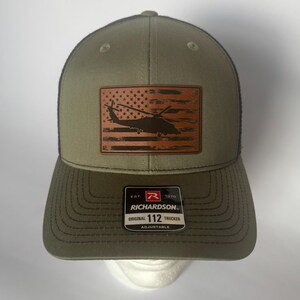 Puede incluir: Gorra de camionero verde oliva con un parche de cuero marrón con un diseño de bandera estadounidense y una silueta de helicóptero. La gorra tiene una parte trasera de malla negra y una visera curva. La etiqueta dice "RICHARDSON ORIGINAL 112 TRUCKER ADJUSTABLE."