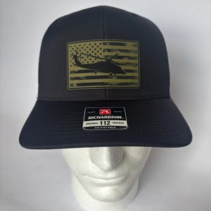 Puede incluir: Gorra de camionero negra con un parche con un diseño de bandera estadounidense y una silueta de helicóptero. La gorra tiene una visera curva y una correa ajustable. El parche es de color verde oliva. El texto "RICHARDSON" y "112 TRUCKER" es visible.