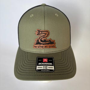 May include: Olive green and black trucker hat with a brown leather patch featuring a snake and the text "no step on snek." The hat has a curved brim and an adjustable strap. The Richardson 112 Trucker hat is adjustable.