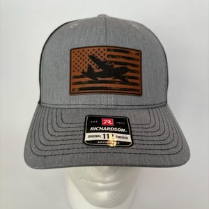 May include: Gray trucker hat with a black mesh back and a curved brim. The front features a brown leather patch with a silhouette of a plane over a distressed American flag design. The hat includes the text "RICHARDSON" and "ORIGINAL 112 TRUCKER".