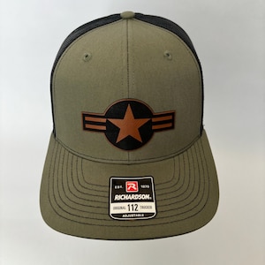 Op de afbeelding: Een groene en zwarte trucker-pet met een bruine leren ster-patch en de tekst "EST. 1970 RICHARDSON. ORIGINAL 112 TRUCKER ADJUSTABLE" op de achterkant.