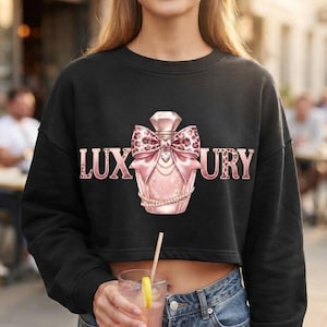 Può includere: Una felpa nera a maniche lunghe e taglio corto con la grafica di un flacone di profumo rosa con un fiocco leopardato e la scritta LUXURY in caratteri leopardati rosa sul petto, che include un dettaglio a forma di collana di perle.