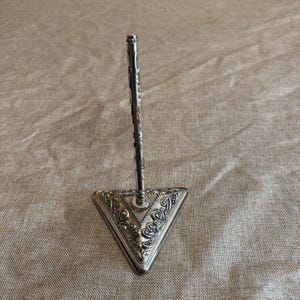 May include: A silver-toned metal stand featuring a triangular base with intricate embossed scrollwork patterns and a single vertical rod extending upward from the center, designed to hold an object upright in a stable position.