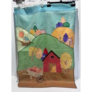 May include: A rectangular fabric wall hanging featuring a landscape scene with rolling green hills, a red barn, and a wheelbarrow filled with orange pumpkins. The design includes colorful appliqued trees in shades of yellow, purple, and pink.