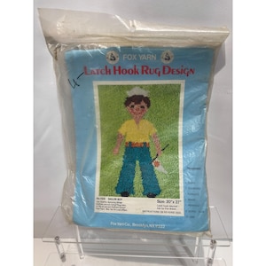 May include: A Fox Yarn latch hook rug design kit in a clear plastic bag. The package features a graphic of a sailor boy on a green background, wearing a yellow shirt and blue pants. The text reads Sailor Boy and indicates a size of 20 inches by 27 inches.