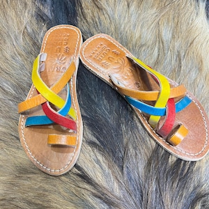 K&ouml;nnte beinhalten: Ein Paar handgefertigte Ledersandalen mit bunten Riemen in Gelb, Blau, Rot und Orange. Die Sandalen haben eine Zehenschlaufe und sind an den R&auml;ndern gen&auml;ht. Die Sandalen werden auf einer strukturierten, braunen Oberfl&auml;che pr&auml;sentiert.