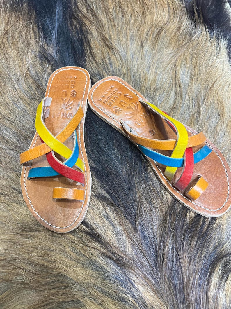 K&ouml;nnte beinhalten: Ein Paar handgefertigte Ledersandalen mit bunten Riemen in Gelb, Blau, Rot und Orange. Die Sandalen haben eine Zehenschlaufe und sind an den R&auml;ndern gen&auml;ht. Die Sandalen werden auf einer strukturierten, braunen Oberfl&auml;che pr&auml;sentiert.