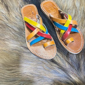 K&ouml;nnte beinhalten: Ein Paar handgefertigte Ledersandalen mit bunten Riemen. Die Sandalen haben eine braune Ledersohle mit Zehenschlaufe und gekreuzten Riemen in Gelb, Rot, Blau und Orange. Die Sandalen sind mit wei&szlig;em Faden gen&auml;ht.